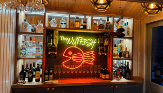Fully stocked garden bar with illuminated shelving, hanging glasses and a neon sign reading &ldquo;The Nutfish&rdquo;.